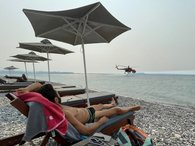 A man sunbathes on a chaise longue on the beach while a helicopter is refueling in the background.