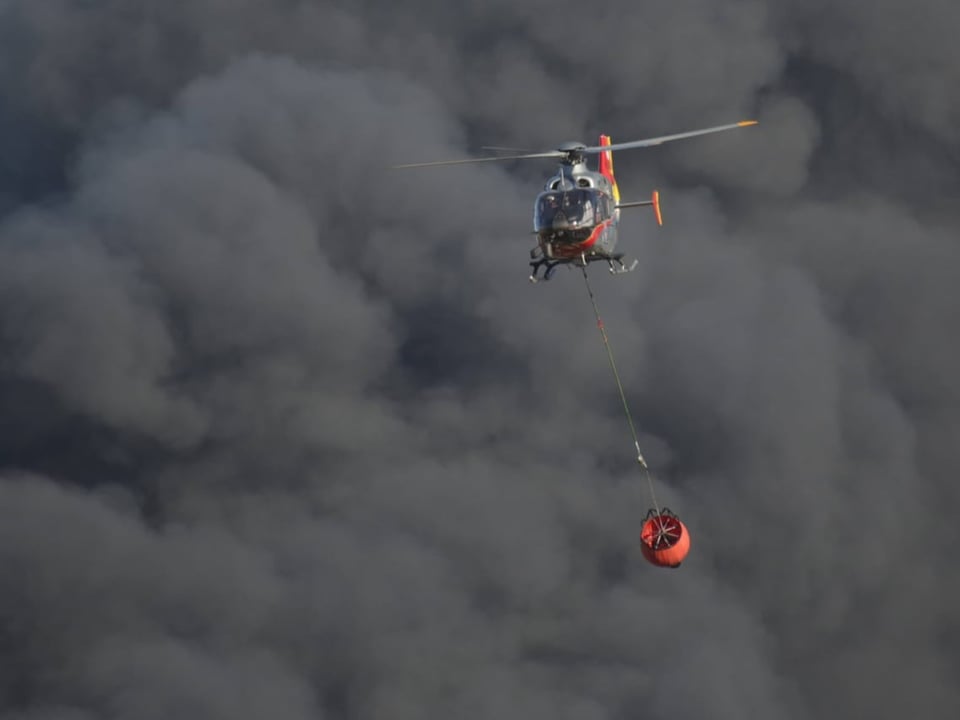 A helicopter carrying water containers amid a thick cloud of smoke