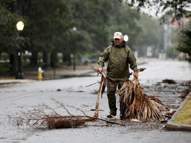 A man is walking across a street carrying branches that have fallen from there.